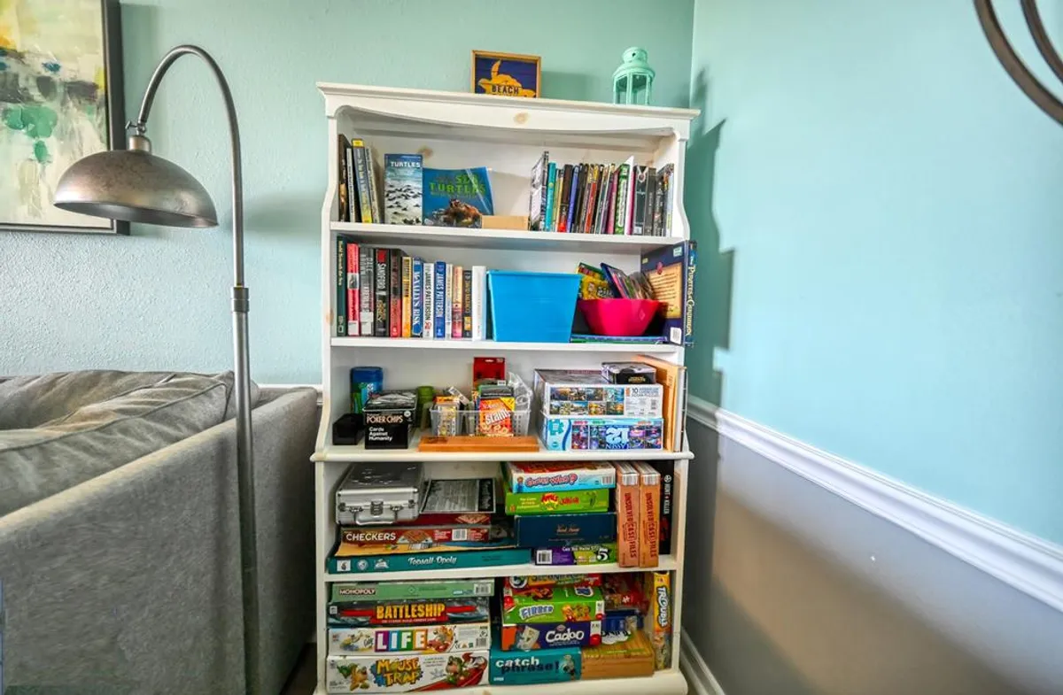 A cozy corner featuring a bookshelf neatly stocked with books and various board games, accompanied by a colorful storage bin and a warm lamp. The setup suggests a family-friendly environment, perfect for gathering after a day of exploration.