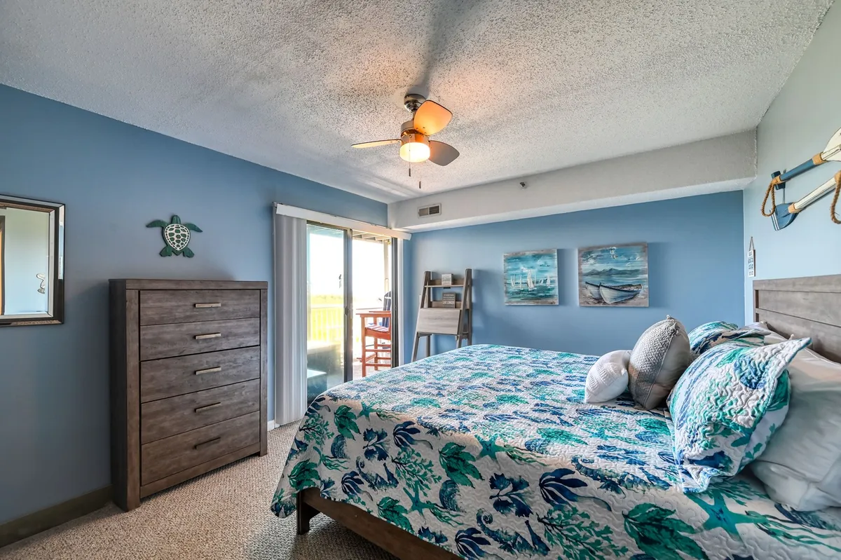 A serene bedroom featuring a comfortable bed with floral bedding, a dresser, tasteful wall art, and a sliding glass door that opens to what appears to be a private balcony. Soft, natural light fills the room, creating a tranquil and inviting space ideal for relaxation.