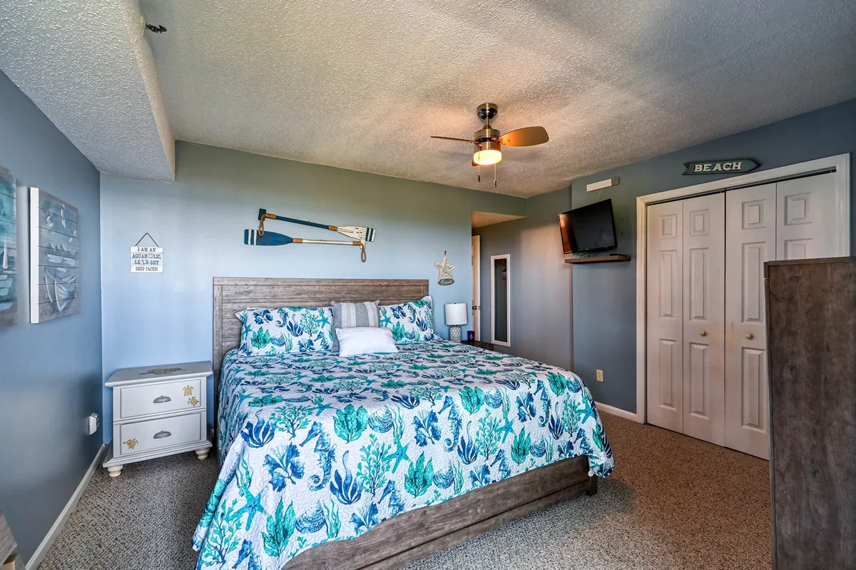 A cozy king bedroom suite featuring floral bedding, a ceiling fan, and nautical decor, with a TV mounted on the wall. The room exudes a calm and inviting atmosphere, suggesting restful mornings with gentle coastal light and moments of relaxation after a day by the ocean.
