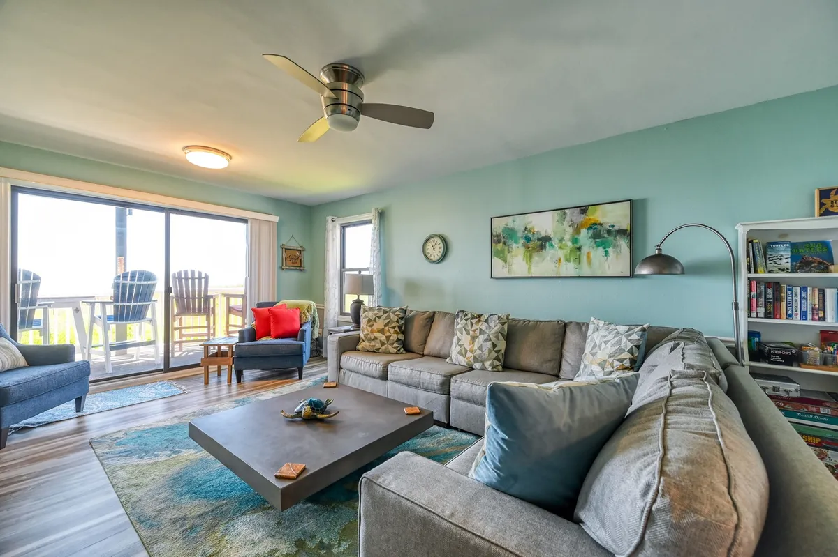 This image captures the spacious, open-concept living room with a large sectional sofa, a coffee table perfect for board games, tasteful artwork, and a sliding glass door that leads directly to the balcony. Natural light fills the space, and the inviting arrangement encourages relaxation and socializing.