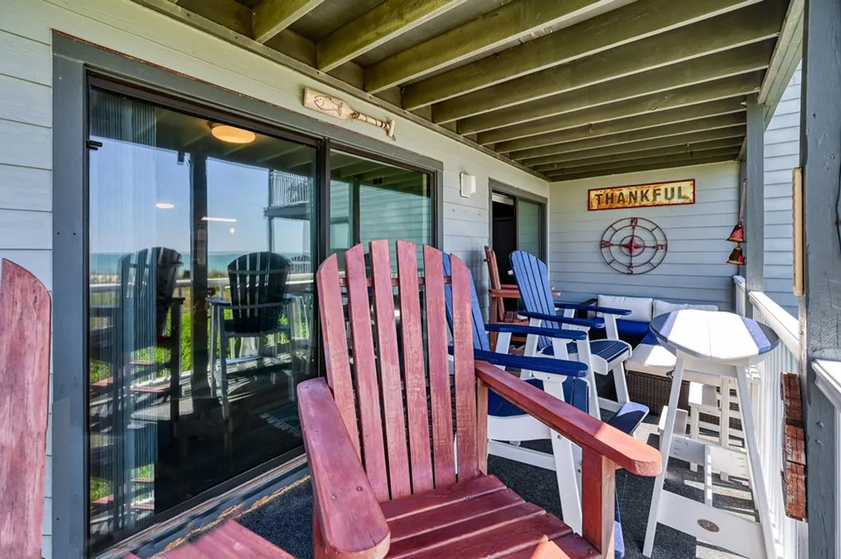The hero image features a welcoming exterior scene with a row of colorful Adirondack chairs facing the ocean, a cheerful 'thankful' sign, and charming nautical decor. The setup invites guests to relax, soak in coastal views, and enjoy conversations with loved ones in a vibrant, seaside atmosphere.