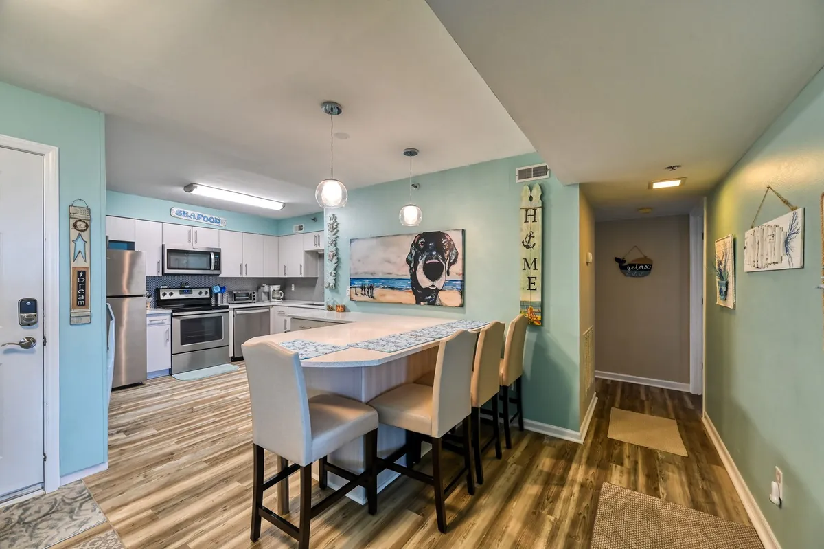 The image features a cozy and inviting kitchen with a prominent kitchen island, modern light fixtures, and a touch of playful decor including artwork of a dog. The space is designed for cooking, family gatherings, and entertaining, with enough room for socializing around the island.