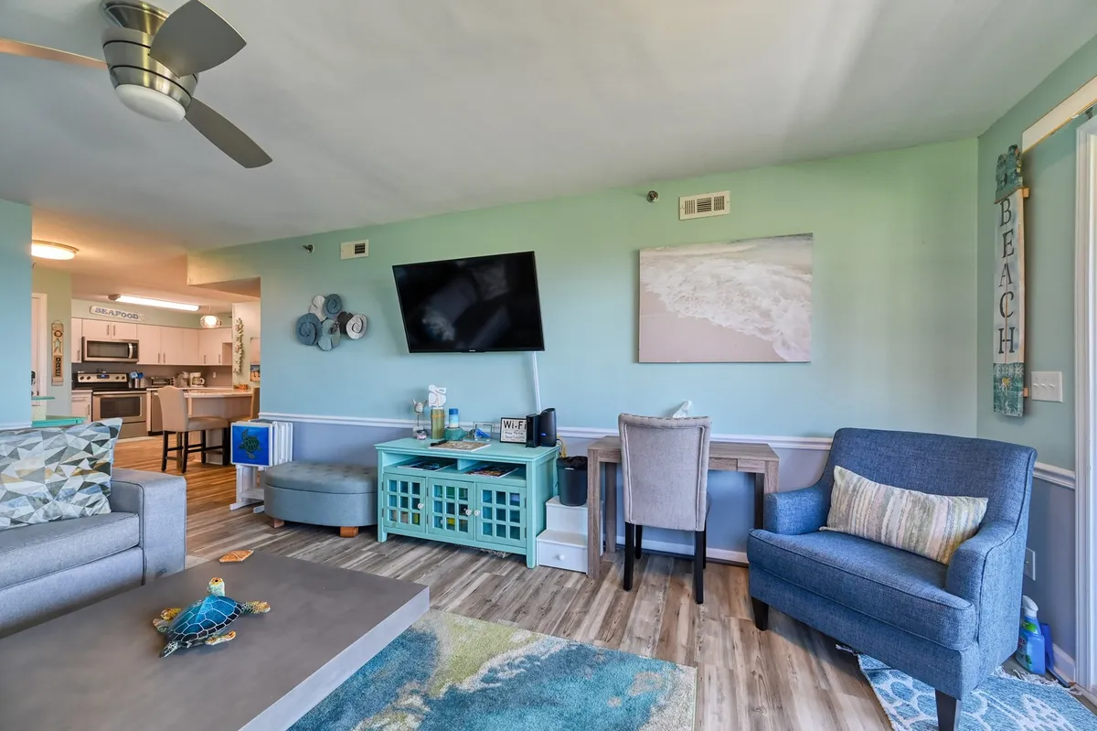 A cozy living room scene with a plush sofa, coffee table set for games or snacks, a TV for movie nights, casual wall art, and a convenient desk area for quick updates or postcards. The room glows with natural light, inviting guests to relax together.