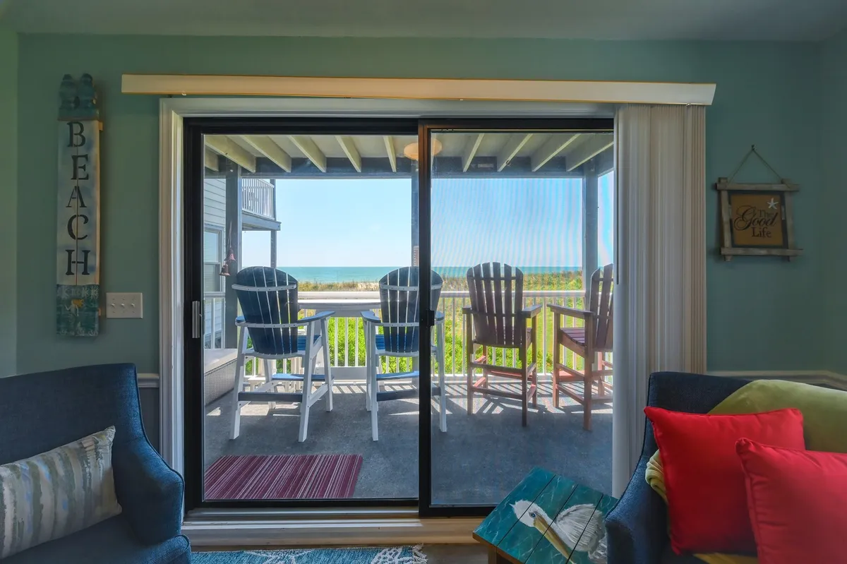 A cheerful and tranquil view through the sliding glass doors to the oceanfront porch, which features Adirondack chairs and a cushioned settee. The porch invites relaxation with panoramic ocean views and beach-themed décor, suggesting sunrise coffees and sunset conversations.