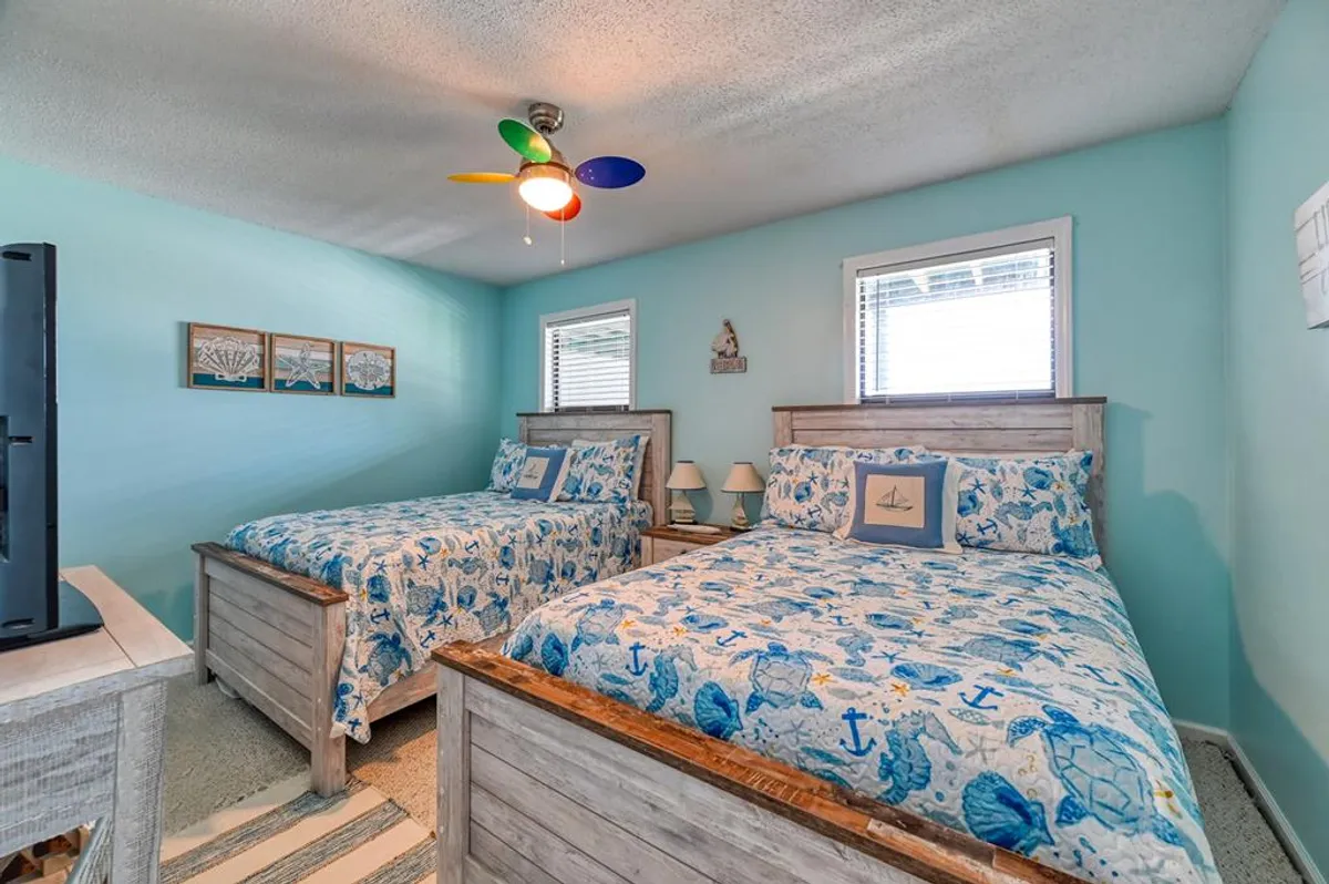 A cheerful interior view with two beds featuring nautical-themed bedding, a colorful ceiling fan, and framed artwork. The space feels playful and relaxed, perfect for friends or couples to unwind after exploring local attractions.