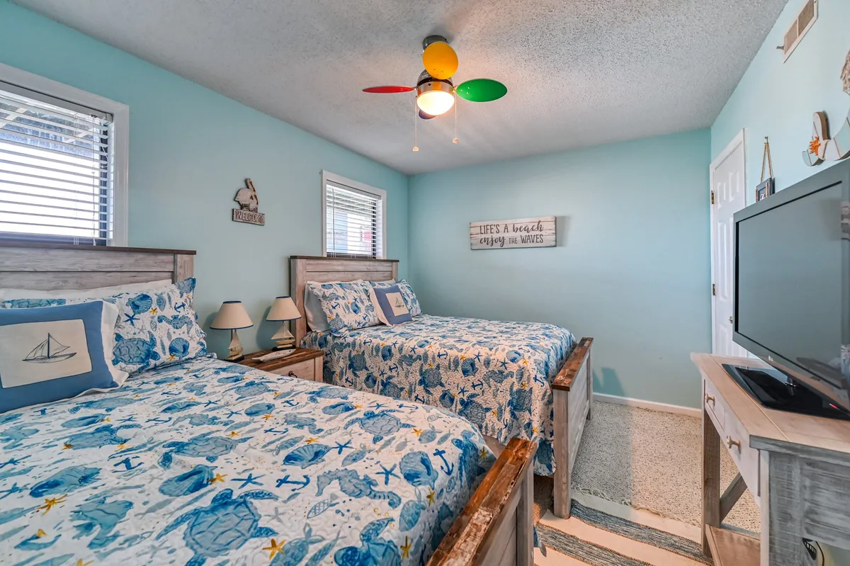 A bright, playful guest bedroom with two double beds adorned in ocean-themed bedding, a colorful ceiling fan, and a cheerful decorative wall sign. The space radiates warmth and is perfectly suited for families or friends sharing the room.