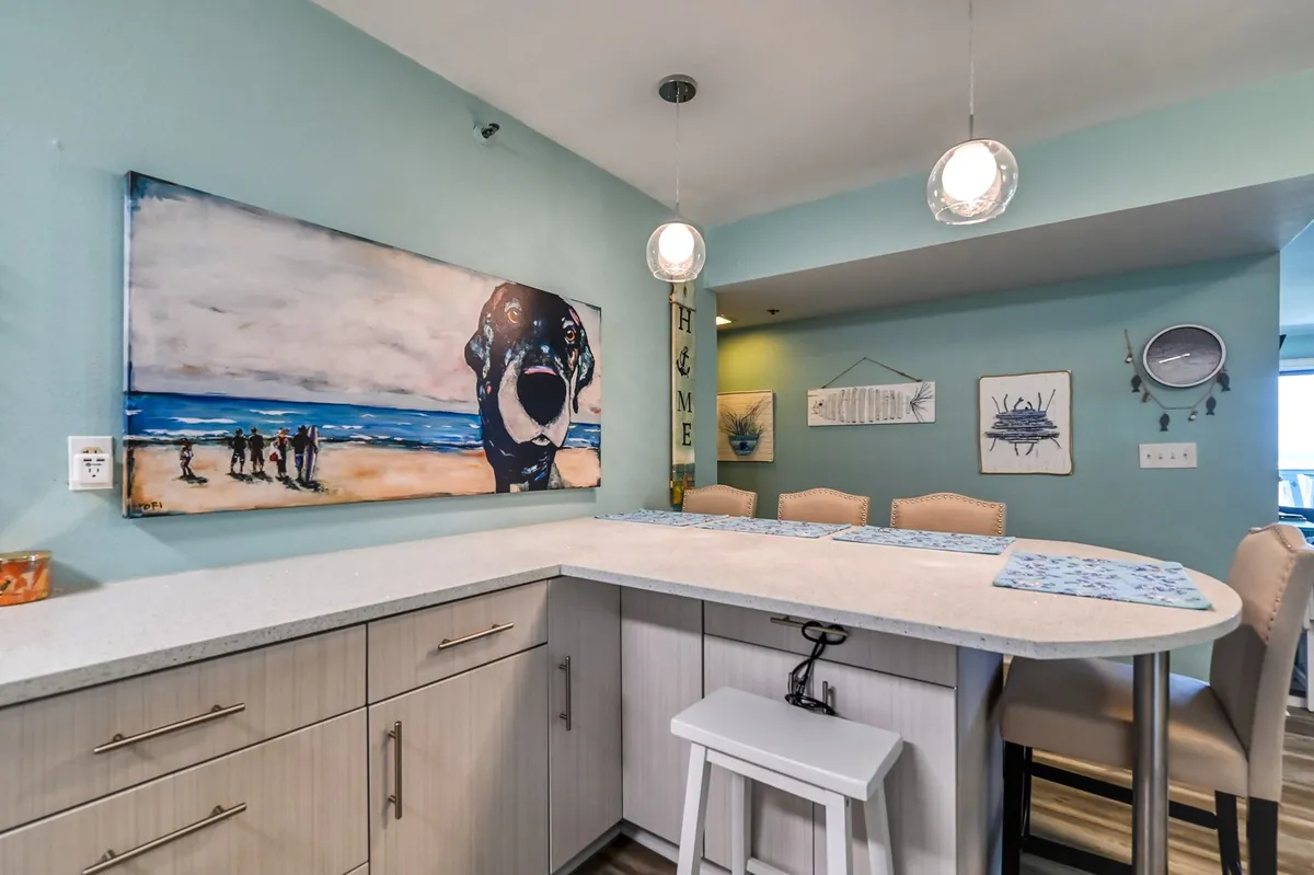 A bright, inviting dining area with a large bar counter, playful wall decor (including a dog artwork), and ample space for socializing. The setup hints at casual gatherings, shared meals, and a sense of community.