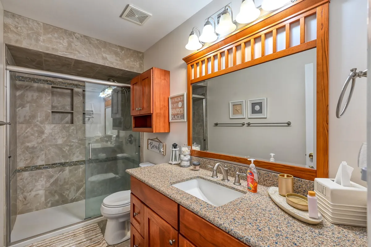 A modern bathroom with a granite countertop, wooden cabinetry, a large mirror, and a glass shower area. The ambiance is clean and calming, ideal for self-care and post-beach relaxation.